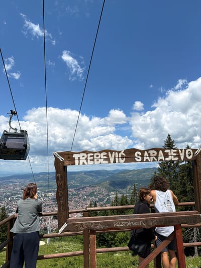 In Sarajevo, Bosnia and Herzegovina, a cable car ascends towards Trebević mountain, passing over a sprawling cityscape. The cable car, labeled with the number "25," is suspended by thick cables against a backdrop of blue sky dotted with white clouds. Below, a wooden sign frames a panoramic view, bearing the inscription "TREBEVIĆ SARAJEVO" in white lettering. Several people are present at the viewpoint. A woman in a grey jacket and dark pants stands with her back to the camera, looking out at the city. Further along the wooden railing, two individuals are embracing: a woman in a black jacket and a man in a white tank top with a graphic print. They stand close together, looking out at the view. The foreground features a wooden fence and greenery, suggesting a scenic overlook. The overall scene captures a moment of appreciation for the urban and natural landscape from a vantage point on Trebević mountain.