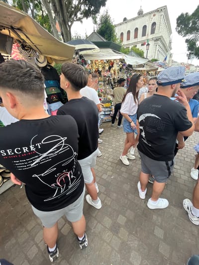 In Venice, Italy, pedestrians are walking along a cobblestone pathway. The scene is bustling with activity as people browse through market stalls. A tall, white, multi-story building with a rounded turret on top stands in the background, adding to the architectural charm of the city. A woman in a white t-shirt and jean skirt is visible, and the surrounding area is filled with the colors and textures of various market goods. The atmosphere is lively, reflecting the typical commerce in a tourist hub of Venice.
