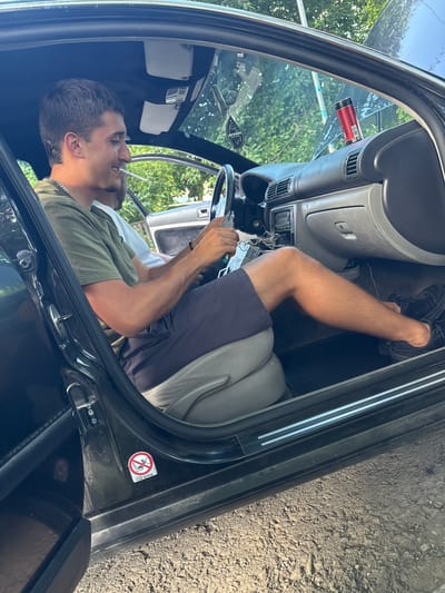 A young man with short, dark hair is sitting in the driver's seat of a dark-colored car, with the driver's side door open. He is wearing a green t-shirt and dark shorts. He is smiling and holding a cigarette in his mouth. His right arm is resting on the steering wheel, and his hands are near the car's dashboard, which appears to be partially disassembled. Another person, whose face is not visible, is seated in the passenger seat. The car's interior is visible, including the dashboard, seats, and a red can on the dashboard. Outside the car, there are trees and foliage, suggesting an outdoor, possibly rural or suburban, setting. The ground outside the car is rough and appears to be gravel or dirt. A sticker on the inside of the car door shows an airbag symbol with a prohibition sign. The location is Topoli, Bulgaria.
