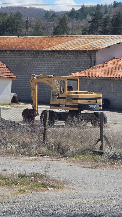 The image presents a "Schaeff HML 30" excavator parked on the edge of a gravel road in Slatino, Bulgaria. The excavator is yellow and black and is located adjacent to a building with a combination of rusted metal and tiled roofing, along with concrete block walls. Beyond the building is a hilly, forested area under a partly cloudy sky. A chain-link fence separates the excavator from the gravel road.