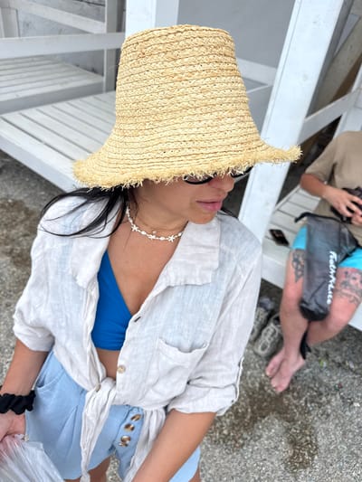 The image shows two people sitting on a white wooden bench outdoors. The person in the foreground is wearing a straw hat, sunglasses, a light-colored open shirt, and a blue bikini top. They are also wearing a delicate necklace with a star charm. The person's skin is visible on their arms and chest.
The person in the background is partially visible, wearing a tan shirt and blue shorts. They are holding a black bag with the text "Featherlite" printed on it. The background shows a rocky or gravelly ground, suggesting an outdoor setting possibly near a beach or waterfront. The lighting suggests it is daytime. The overall mood appears relaxed and casual.