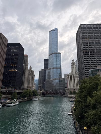 The image captures a view of the Chicago River and the city's skyline. In the foreground, the murky green water of the river flows. Along the riverbanks, several tour boats are docked or moving. On the left side, a white and green tour boat with "CITY LIGHTS" written on it is visible. Further down, another similar boat is docked. On the right bank, trees line the riverwalk, with a few boats visible further up.
Dominating the skyline are numerous skyscrapers. The most prominent structure is the Trump International Hotel and Tower, a tall, glass-covered building with a distinctive spire. To its left, another tall, slender skyscraper with a pointed top stands out. Various other buildings of different architectural styles and heights fill the background, creating a dense urban landscape. Some of the buildings have a grid-like facade, while others feature more decorative elements.
The sky is overcast with heavy clouds, casting a diffused light over the scene and giving it a somewhat subdued atmosphere. Bridges span the river in the distance, with traffic visible on them. The overall impression is that of a bustling city on a cloudy day, with the river acting as a central artery through the urban environment.