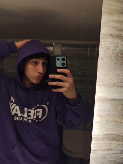 A young man takes a selfie in a bathroom mirror. He wears a purple hooded sweatshirt with a white logo that reads "RELAY FOR LIFE" in reverse. His face is slightly puckered, and he holds a black smartphone with a teal camera area. The mirror shows a dark, possibly tiled wall behind him. The edge of the mirror reveals a textured, light-colored wall.
