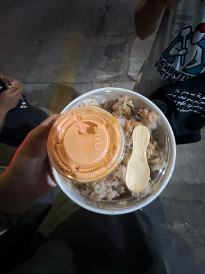 An overhead, dimly lit image captures a person's hand holding a clear plastic food container in León, Mexico. The container holds a mixed rice dish, topped with a small plastic cup featuring an orange lid labeled "SOLO" and a small wooden spoon. To the left, the arm and lap of a seated individual wearing dark pants and a white shirt are visible. To the right, another person's torso is seen, wearing a light-colored t-shirt with a blue and white graphic depicting two figures holding tools, and the text "CEMENTO FORTAL". The ground beneath is a dark, textured paved surface, possibly a sidewalk, with a faint yellow marking. The scene appears to be taking place at night or in a low-light indoor setting.
