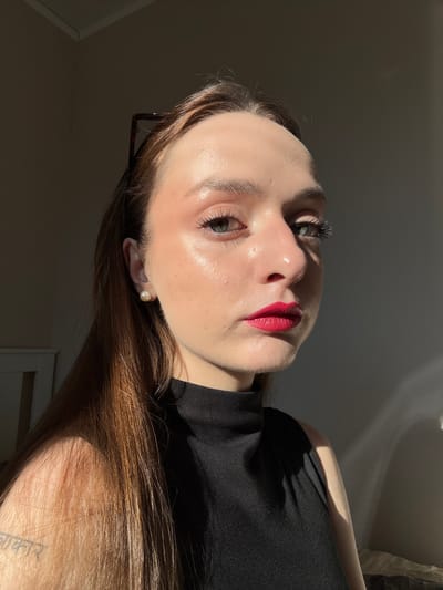 A young woman with long, light brown hair is positioned in a medium close-up shot, looking towards the right of the frame. She is wearing a black, sleeveless, turtleneck top. Her face is illuminated by a strong side light, creating a noticeable highlight on her cheek and nose. She has bright green eyes, well-defined eyebrows, and prominent eyelashes. Her lips are painted with a vibrant red lipstick. A single pearl earring is visible in her left ear. There is a tattoo on her left shoulder blade. The background is dimly lit, with a shadow cast on the right side of the frame. The overall impression is that the photograph was taken indoors, likely in a room with natural light. There are no visible indicators to suggest a specific location within Palilula Urban Municipality, Serbia.