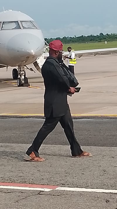 A person in a black suit and a red cap walks on an airport tarmac near an airplane. The person is carrying a dark rectangular object in their right hand. Another person in a yellow vest stands in the background. The location is the Orerokpe Airport in Orerokpe, Nigeria. The sky is cloudy.