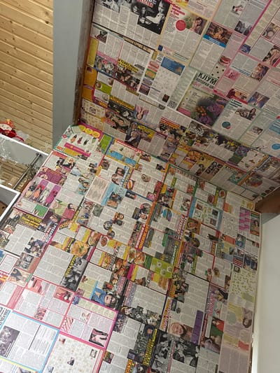 This image displays a room where the walls and ceiling are covered with a collage of various newspapers and magazine pages. The pages are layered and overlapping, creating a dense mosaic of text and images. The content of the pages appears to be varied, including photographs of people, articles, advertisements, and what seems to be puzzles or games. Some pages feature close-ups of individuals, while others show group photos or larger scenes. There are also images of food, products, and landscapes. The text is in Russian, with headlines and articles visible throughout the collage. Some headlines include "КЖУМ! всё лето" (KZhUM! all summer), "Купила себе ДЕРЕВНЮ!" (Bought myself a VILLAGE!), "С ребенком ктожтей!" (With a child who is stronger!), and "Из России на п приехал только" (From Russia to n only arrived). The overall impression is that of a space decorated with a personal collection of printed media, perhaps a creative or unconventional interior design choice. The lighting suggests it is daytime, and the setting appears to be indoors, possibly a living space or a creative studio. The tone is eclectic and visually busy due to the sheer volume of printed material covering every available surface.
