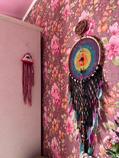 The image captures a close-up view of a dreamcatcher hanging from a floral-patterned ceiling in Krasnoe-na-Volge, Russia. The dreamcatcher has a circular woven center with concentric rings of vibrant colors, including purple, blue, yellow, and orange, and is adorned with feathers in various pastel shades and small red beads. A wooden ornament with intricate carvings is attached to the left side of the dreamcatcher's hoop. Below the dreamcatcher, a festive hanging decoration with pink fringe and a triangular fabric piece featuring a UFO illustration is visible. The setting appears to be indoors, likely a room, with soft pink walls and a portion of a wooden shelf visible. The lighting suggests daytime, and the overall mood is whimsical and decorative. No visible text is present in the image.