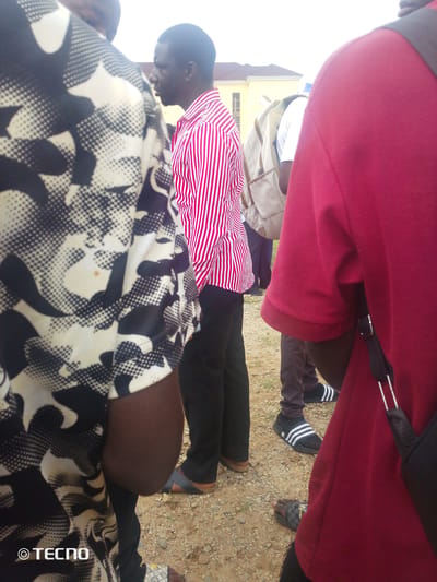 In Gumel, Nigeria, a group of individuals stands on a dirt ground. The person in the foreground, seen from the back, wears a red shirt and a black bag with silver straps. To their left, a person in a black and white patterned shirt is partially visible, with their arm exposed. Further left, a man in a red and white striped shirt faces to the right, his left side visible. He wears black pants and dark sandals. Behind him, another individual in a white shirt and a beige backpack is also present. A building with a yellow facade and a tiled roof is visible in the background.