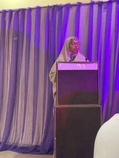 A person wearing a hijab and glasses is speaking into a microphone in front of a backdrop of purple and grey striped fabric. The person is positioned to the left of two large, dark rectangular objects, likely speakers or monitors. The backdrop is illuminated with purple lighting, creating a stage-like atmosphere. The person appears to be presenting or speaking at an event. The scene is indoors, with no indication of the time of day or weather conditions. There is no visible text in the image. The location context is Unguwar Shanu, Nigeria.
