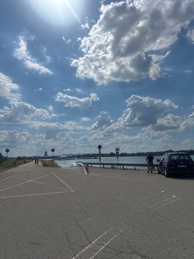 A vertical image depicts a sunny day at a waterfront. The sky is a vibrant blue, filled with large, puffy white clouds that are brightly illuminated by the sun, which is visible in the lower left corner as a lens flare. The scene appears to be a paved area, likely a parking lot, with white lines marking parking spaces.
In the background, a body of water stretches horizontally across the frame, reflecting the sunlight. Along the edge of the water, a metal railing separates it from the paved area. Beyond the water, a distant shoreline is visible with trees and some structures, possibly a marina with sailboats.
On the paved area, near the water's edge, a dark-colored car is parked. A person, dressed in a dark t-shirt and shorts, stands beside the car, facing away from the camera and towards the water. Another person, also dressed in casual clothing, is further down the paved path, seemingly walking or standing. Several street signs and poles are also present along the path.
The overall atmosphere is one of a leisurely day outdoors, possibly at a park or recreational area by a lake or river in Altenheim, Germany. The weather is clear and sunny, suggesting it is daytime. The visible license plate on the car reads "GM-074-RE".