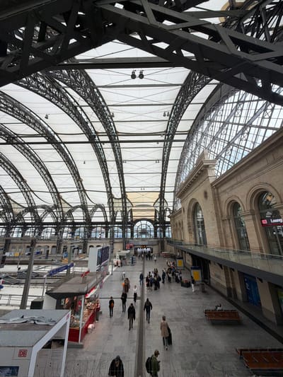 This is an interior shot of Dresden Hauptbahnhof (Dresden Central Station) in Dresden, Germany. The architectural design features a large, arched ceiling with exposed steel framework and white panels. Below the ceiling, the train station platform is visible with people walking, standing, and waiting. There are kiosks and signage visible on the platform. The foreground shows structural elements of the station's framework.