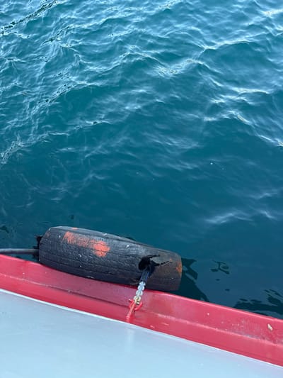 In Lepetane, Montenegro, a close-up view of a boat's edge shows a worn, black fender, possibly a repurposed tire with orange markings, attached to the red-painted hull with a chain. The fender floats in dark blue, rippling water, reflecting the sky and creating a sense of movement. The boat appears to be docked or moving slowly, with the focus on the functional and slightly weathered protective element against the natural backdrop of the Adriatic Sea.