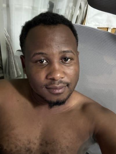 The media file is a close-up, low-angle selfie of a man in what appears to be a room, possibly at home or a hotel, given the presence of a gray, textured surface that resembles a headboard or furniture upholstery, and a stack of folded towels or linens in the background. The man is of African descent, with dark, curly hair and a short beard and mustache. He is looking directly at the camera with a neutral expression. His chest and shoulder are visible, showing some body hair. The lighting appears to be artificial and somewhat dim, suggesting it might be nighttime or indoors with limited natural light. There is no visible text in the image. The orientation of the image is such that the man's head is to the left and his body extends to the right, suggesting he took the photo by holding the camera at an arm's length above him and to his side. The overall impression is a casual, personal snapshot.