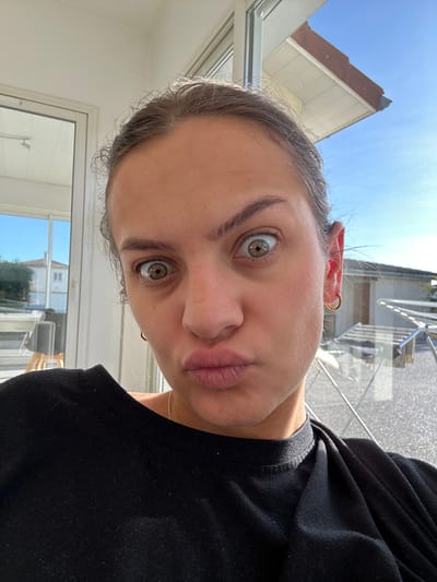 A close-up image depicts a young adult female located indoors in Veauchette, France. The female subject, positioned centrally, has fair skin, visible brown hair pulled back, wide-open hazel eyes, and her lips are pursed forward. She is wearing a dark, possibly black, long-sleeved garment and gold hoop earrings in both earlobes.
The background, visible through what appears to be a glass door or large windows, reveals an outdoor residential scene under a clear blue sky. To the subject's left, a white building with a red-tiled roof and green foliage is visible in the distance. To the subject's right, an outdoor rotary clothes drying rack with a white frame is visible on a light-colored paved surface, beneath a building eave with brown roof tiles. The overall lighting suggests a bright, sunny day.
