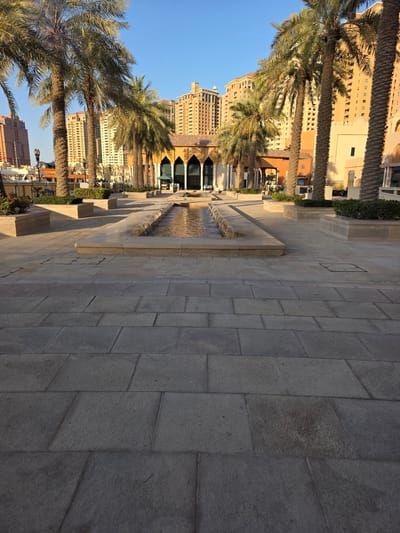 This image captures a sunlit outdoor scene with palm trees and modern buildings in Doha, Qatar. The foreground features a wide pathway paved with large, rectangular stone tiles, some of which show signs of wear or staining. A shallow, rectangular fountain with water jets is centrally located in the mid-ground. Flanking the fountain are raised planters with manicured green shrubs.
Beyond the fountain, a series of tall date palm trees with fronds casting shadows on the ground are prominent. Behind the trees, a light-colored building with arched windows and intricate architectural details is visible. Further in the background, several multi-story modern buildings with light-colored facades and numerous windows rise against a clear blue sky. The architectural style of the buildings suggests a contemporary development, likely in a tourist or commercial district. No people are visible in the frame. The lighting indicates it is likely late afternoon or early morning, given the long shadows cast by the palm trees. The overall environment appears clean and well-maintained.