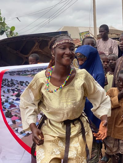 In Rigachikun, Nigeria, a woman with braided hair, wearing a white and gold traditional outfit and a beaded necklace, smiles broadly at the camera. She stands in front of a banner depicting an aerial view of a flooded town. Behind her, a crowd of men, women, and children are gathered, some looking towards the camera, others engaged in conversation. The environment appears to be outdoors, with a cloudy sky and utility lines overhead. A car hood is visible on the left side of the frame.