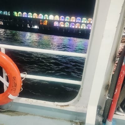 This image captures a nighttime scene on the waterfront in Bhola, Bangladesh, viewed from the deck of a vessel. In the lower-left foreground, an orange lifebuoy with white straps and dark Bengali script (likely "ভোলার") is suspended from a white metal railing. The railing extends horizontally across the lower-mid portion of the frame, separating the viewer's light-blue painted deck from the dark, rippled water. To the right, a white vertical support beam is visible, alongside bundled dark and red pipes or cables. Across the water in the background, a large passenger vessel, possibly a ferry or launch, is brightly illuminated. This vessel displays multiple decks, each featuring numerous arched windows or cabin openings emitting various colored lights (green, blue, purple, yellow, orange), which reflect vividly on the dark water's surface. The sky above the distant vessel is black. No individuals, specific actions, or interactions are discernible.
