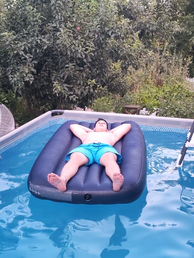A young person is relaxing on a dark blue inflatable mattress in a clear blue swimming pool. They are lying on their back with their hands behind their head and wearing blue swim trunks. The pool is surrounded by a light-colored frame and a decorative blue and white tiled border. Behind the pool, lush green trees and bushes create a natural backdrop. The lighting suggests it is daytime, likely late afternoon or early evening, as the sun is not directly overhead. The overall scene conveys a sense of relaxation and enjoyment on a warm day. There is some visible text on the side of the inflatable mattress, but it is too small and blurry to be legible. The location context provided is Yalangach, Uzbekistan.
