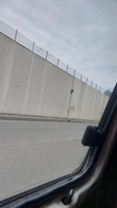 A view from inside a car, through the open window, shows a concrete structure, possibly a dam or a tall wall. It has horizontal metal bars running up its side. The sky is overcast and light gray. A narrow road runs parallel to the concrete wall. The car frame is visible around the open window.