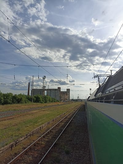 Captured from a moving train, the photograph shows a section of railway tracks. To the left of the tracks, there's a grassy area with remnants of what appears to be an old platform or support structure. In the background, there are industrial buildings, including a tall chimney and lattice towers. Overhead, power lines crisscross the sky. The sky is partially covered with clouds. The train from which the photo is taken occupies the right side of the frame. The location is likely Nova Zagora, Bulgaria.