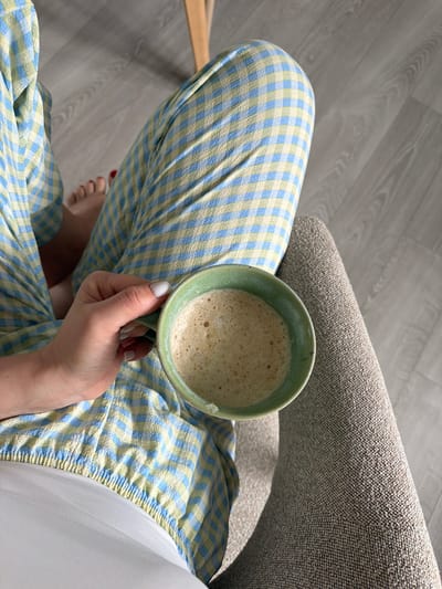 An overhead, close-up shot shows a person sitting in a chair, holding a green mug filled with a frothy beverage. The person is wearing blue and yellow checkered pajama pants and has a white painted fingernail. Their feet, with red painted toenails, are visible in the background. The floor appears to be light wood. The overall scene suggests a casual, domestic setting, likely a home. There is no information in the image that would indicate a location in Dreiliņi, Latvia.