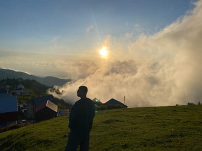 This media file depicts a scenic view of Gomismta, Georgia, likely during the late afternoon or early evening, as indicated by the warm, low-angled sunlight and the presence of clouds illuminated by the sun.
The foreground features a grassy hillside with a person silhouetted against the sky. The person is facing away from the camera, looking out towards the landscape. They are wearing a dark long-sleeved shirt and pants.
In the midground, a small village is nestled on the slopes of rolling hills. Various houses with distinct red roofs are visible, along with some larger structures. The village appears to be situated at a high elevation, with clouds partially obscuring some of the distant hills.
The background is dominated by a dramatic sky filled with luminous clouds. The sun is visible, shining brightly and creating a lens flare effect. The light filtering through the clouds casts a soft, diffused glow over the entire scene, contributing to a peaceful and somewhat ethereal atmosphere. The landscape suggests a mountainous region, characteristic of Gomismta.
There is no visible text in the media file. The overall impression is one of tranquility and natural beauty, capturing a moment of contemplation in a remote, mountainous setting.