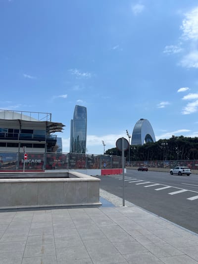 The image captures a street scene in Baku, Azerbaijan, on a clear day with a bright blue sky and scattered clouds. In the foreground, a light-colored stone planter box sits on a tiled pedestrian area. Beyond this, a paved road runs across the scene, marked with white lines indicating lanes. Two cars, one dark red and one white, are visible on the road. A chain-link fence with banners advertising a racing event, possibly Formula 1, runs along the side of the road, featuring images of race cars and drivers. Behind the fence, a multi-story building with a white and dark facade is under construction or renovation. Further in the background, the distinctive modern architecture of Baku is evident, with two tall, glass-covered skyscrapers dominating the skyline. One skyscraper has a sleek, curved design, while the other is more traditionally vertical. A dense line of dark green trees separates the road from the buildings in the distance. Streetlights are visible along the roadside. The overall impression is of a dynamic urban environment with ongoing development and a focus on automotive racing events.