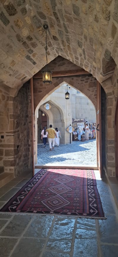 The image shows a view through a stone archway into a bustling outdoor market area in Baku, Azerbaijan. A traditional red and black patterned rug is laid out on the stone-paved floor leading into the arch. Inside the archway, two people are walking away from the camera, followed by another person dressed in white. Further in the background, stalls are visible, with people browsing. The architecture features stone walls and arched doorways, typical of the Old City of Baku. The lighting suggests it is daytime, with sunlight casting shadows on the ground.