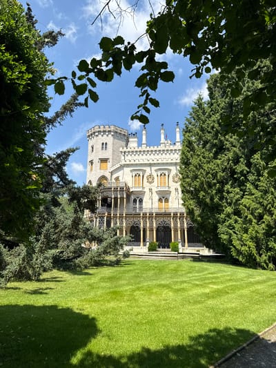 The image depicts Hluboká Castle, a Neo-Gothic landmark located in Hluboká nad Vltavou, Czechia. The castle, built of white stone, features crenellated towers, ornate balconies, and numerous windows. It is framed by lush green trees and a well-maintained lawn, with a bright blue sky dotted with white clouds overhead. The scene is bathed in sunlight, casting shadows on the grass. No people are visible in the image.