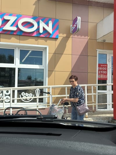 In Valday, Russia, a woman with short brown hair and glasses, wearing a dark blue floral print blouse and blue jeans, walks with a bicycle in front of a building. The building has a facade of alternating beige and pinkish-brown square tiles. A large sign with the letters "OZON" in white 3D lettering against a pink and blue striped background is mounted on the building. To the right of this sign is a smaller, oval-shaped sign also displaying the "OZON" logo. To the right of the woman, a red sign with white text indicates store hours: "08:00" and "21:00" with a "МАГНИТ" logo above. The woman is carrying a purple purse on her left shoulder. She is pushing a bicycle with a metal basket on the front, containing a pink plastic object and other items. A white metal railing with decorative circles and shapes is in the foreground, partially obscuring a window with a reflection of trees and sky. The scene appears to be captured from inside a vehicle, as indicated by the presence of a windshield wiper in the bottom of the frame.