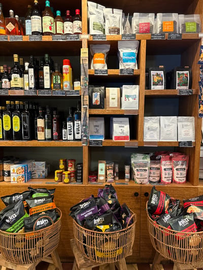 The image depicts a vertically oriented, eye-level view of a shelf display in a shop, likely located in Miraflores, Peru based on the products. The shelves are made of light-colored wood and contain various products.

On the upper shelves are multiple packages of coffee beans with labels such as "Cafe Eight", "Cafe Noos", "Camucha", "Chabela", and "Tarku". Various bags of salt are stacked in multiple layers, including one labeled "Amazon Salt".

The lower shelves contain a selection of bottled items, mostly olive oil, vinegar, and other condiments. Brands like "Colavita", "Kulli", "Casa Rinaldi", and "Carandini" are visible.

Wicker baskets, angled on the right side, overflow with "Inka Chips" in various flavors, including BBQ, Jalapeno, and "Queso & Cebolla". Jars with sardines, tuna, and nuts are placed in the bottom right corner of the frame.

The environment is well-lit and the focus is on the variety of products available for purchase.