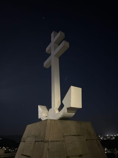At night in Gourock, United Kingdom, a concrete structure featuring a large, illuminated white cross is captured in the lower half of the frame against a dark, starry sky. The cross, comprised of intersecting rectangular sections, extends horizontally from the central base of the structure. The structure itself, partially visible on the right, features layered concrete blocks and several plaque-like elements affixed to its surface. The lighting is muted, emphasizing the stark contrast between the illuminated cross and the dark sky. Distant city lights are visible along the upper right edge of the image.