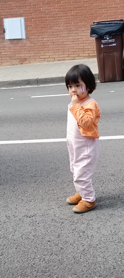 An outdoor, eye-level photograph depicts a young child standing on an asphalt road in Ripollet, Spain. The child has short, dark hair and a light complexion, with a purple and pink butterfly-like face painting on their right cheek. The child wears an orange long-sleeved shirt layered under light pink overalls and tan or brown shoes with velcro closures. Their right hand is raised to their mouth, with fingers touching their lips, and their gaze is directed towards the left side of the frame.

The asphalt road surface is dark gray, marked with a continuous white line in the foreground and a segmented white line further back. A gray concrete curb separates the road from a lighter gray sidewalk. Behind the sidewalk, a reddish-brown brick wall is visible, constructed with a stretcher bond pattern. On the left side of the wall, a rectangular white utility box with a dark latch is mounted. On the right side of the frame, partially obscured by the child, a brown public waste bin is situated on the sidewalk. The bin has a black plastic bag protruding from its top opening and features text, including "ripollet aporta," confirming the location. The scene is illuminated by ambient daylight, with no harsh shadows present.