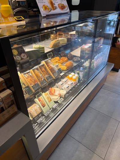 This is a slightly tilted, medium-angle shot of a glass display case filled with various food and beverage items, likely located in a cafe or bakery setting. The case contains a variety of pastries, sandwiches, salads, and drinks, neatly arranged on shelves. The camera angle reveals some of the items from the front and top. Signs and labels are visible, some with Chinese text. The floor is made of large grey tiles, and wooden elements are visible along the edges of the display case. The location may be within the city of Yuhuatai District, China, if that information is needed to provide context.