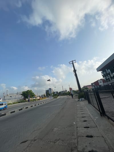 The image depicts a street scene in Ilado, Nigeria. A gray concrete road runs horizontally, bordered by a gray curb with black rectangular markers. A building with a modern, possibly partially reflective facade is visible in the upper left corner. A sidewalk with a black metal fence runs along the upper right edge of the road. In the background, the sky is blue with scattered white clouds. An electrical crane stands to the left of the road, with power lines strung to a pole on the right side of the road. Several buildings, trees, billboards, and a blue vehicle are visible further down the road, along with pedestrian figures on the roadside.