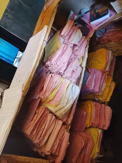 The image shows a disorganized pile of papers, predominantly pink and yellow, stuffed into wooden containers. The papers appear to be documents or records, some with handwritten text visible. One stack of papers has a book titled "HON FROM THE ROCK" perched on top. Another stack features a bottle of water. In the background, a blue translucent sheet is visible, possibly a window or partition. The setting seems to be an indoor storage area, possibly an office or a storeroom in Nigeria, given the location context. The lighting suggests it could be daytime, but the overall impression is one of clutter and neglect, with no clear activity taking place beyond the storage of these materials. Text visible on some papers includes "Tai Albay", "CLA", "NAKOWA CUNG& TERNITY", "ARLEY", and "INE INJECTION".