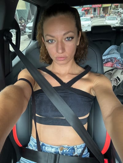 A woman is seated in a car wearing a black top and denim shorts. A seatbelt is across her chest. Outside the car window appears to be a busy street scene in Sa Pa Ward, Vietnam, with parked vehicles and buildings visible. There are garments on the back seat. The image is taken from a close, angled perspective from above.