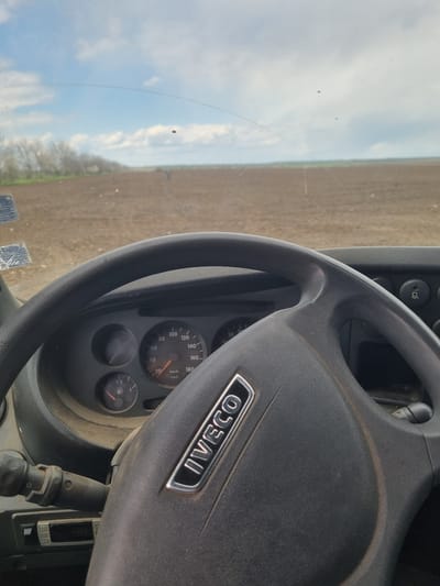 The image shows the interior of an IVECO vehicle, specifically the steering wheel and dashboard. The steering wheel is black with the IVECO logo displayed. The speedometer indicates a speed in kilometers per hour, and a fuel gauge is visible. Outside the vehicle, a field and sky can be seen through the windshield. The image does not provide sufficient information to determine the exact location within Dobrevo, Bulgaria, or include details about people, actions, or interactions.