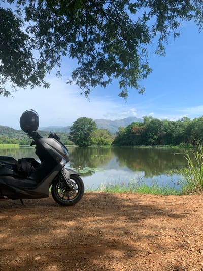 A dark grey scooter is parked on a dirt path next to a calm lake. A black helmet rests on the handlebars of the scooter, and a dark fabric item is draped over the seat. The lake is surrounded by lush green trees and rolling hills in the distance. The sky is a clear blue with a few scattered clouds. The overall scene suggests a peaceful, natural setting, likely in a rural area. The location is the city of Buduruwagala, Sri Lanka.