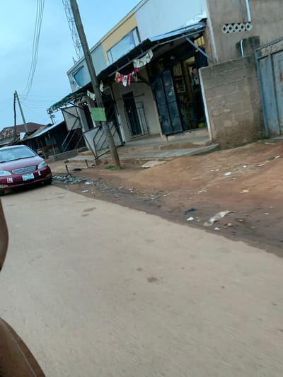 The image depicts a street scene in Du, Nigeria. A maroon sedan is parked on the left side of a paved road. The car has a visible license plate that reads "BAA 196 AE". Beyond the road, a building with a light-colored facade and a dark awning stands. Various items, including what appear to be flags or decorations, are hanging from the awning. A person is partially visible inside the building through an open doorway. Adjacent to the building, a wall constructed of concrete blocks is visible, with a metal gate to its right. The ground area between the road and the building is unpaved and appears to be a mixture of dirt and debris. Electrical wires are suspended overhead. The overall environment suggests a commercial or residential street in a town or city.