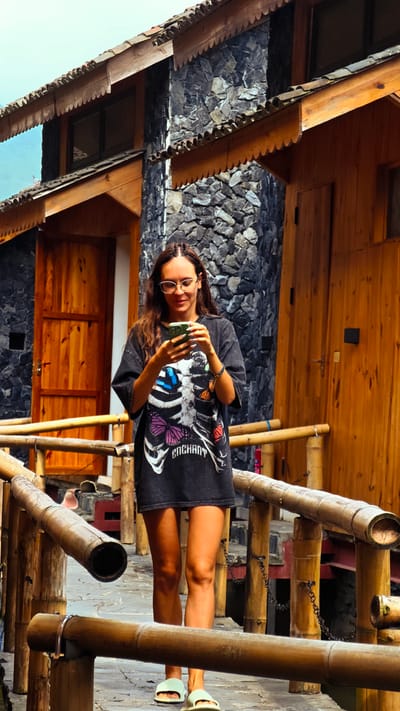 A young woman stands on a bamboo walkway, focused on her smartphone, in Du Già Commune, Vietnam. She has long brown hair, partially braided, and wears round glasses, looking down at her device with a subtle smile. Her casual attire includes a loose, dark grey oversized T-shirt featuring a graphic of a white skeletal rib cage embellished with colorful butterflies in shades of blue, purple, and orange. The word "ENCHANT" is visible on the lower part of the design. She completes her look with light green, open-toed slide sandals.

Behind her are traditional buildings characterized by dark grey, stacked stone walls and wooden structures, including closed doors, window frames, and decorative roof eaves with serrated edges. The walkway she stands on is made of concrete or stone, flanked by rustic bamboo railings, some sections connected by chains. The sky above is a clear blue, indicating bright daylight. The scene suggests a tranquil outdoor setting, possibly a village or resort, blending natural materials with traditional architecture.