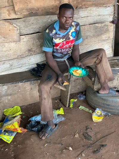 In Gbeleko, Nigeria, a dark-skinned man is sitting outdoors, possibly on or near a wooden platform, appearing to eat. He's wearing a multicolored camouflage t-shirt with "Napoli" written on it, brown pants, and black sandals. He is holding a green plastic plate with food and a piece of orange fruit in his hand. His right foot rests on a tire. Litter, including yellow plastic bags, is scattered on the ground. The ground appears to be dirt and mud.
