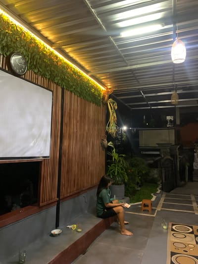 A person is lying on the ground, holding a phone and appearing to be engrossed in it. The setting is an outdoor covered area with a corrugated metal roof and wooden walls. There are plants, including ferns, growing along the wall. A clock is visible on the wall. The lighting suggests it is nighttime, with warm yellow lights illuminating the plants and the area. In the background, there's a glimpse of what appears to be a traditional Indonesian building structure. The overall scene conveys a relaxed and tranquil atmosphere.
