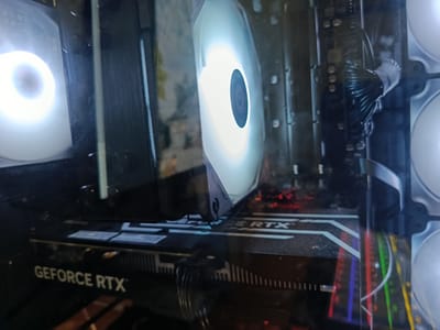 The image shows the interior of a computer case, likely a gaming PC, viewed from an angle. The primary focus is on a large, white-lit CPU cooler fan, which is prominent in the center of the frame. To the left, another fan with a similar white illumination is visible. A graphics card, identified by the "GEFORCE RTX" branding on its shroud, is mounted horizontally in the lower portion of the case. Behind the graphics card and CPU cooler, various internal components of the computer are visible, including cables and what appears to be motherboard elements. The lighting is dominated by the internal RGB lighting of the fans, creating a bright, diffused glow. Reflections are present on the computer case's transparent side panel, obscuring some details of the environment. The context of Ust-Ilimsk, Russia, is not discernible from the visual information provided in the image.