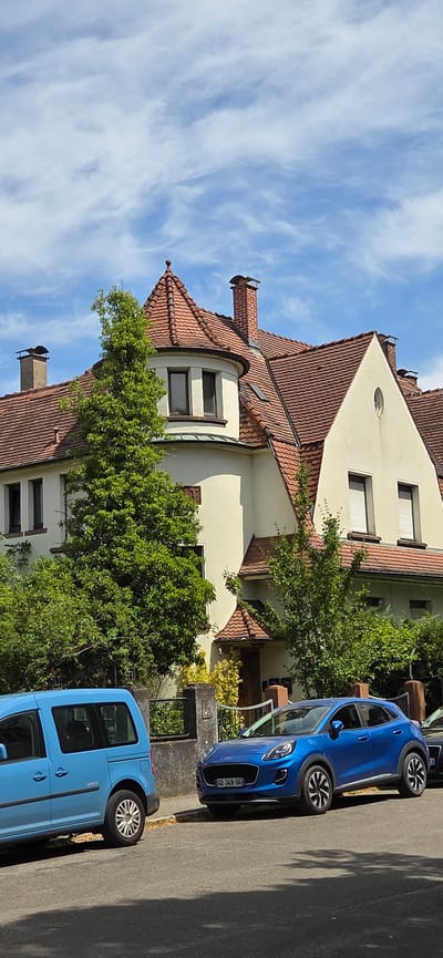 A bright blue Ford Puma is parked on a residential street in Kehl, Germany, next to a bright blue van. The cars are parked in front of a large, two-story house with a red-tiled roof and cream-colored walls. The house has a prominent round turret with a pointed roof. Mature trees and greenery surround the house. The sky is blue with scattered white clouds.