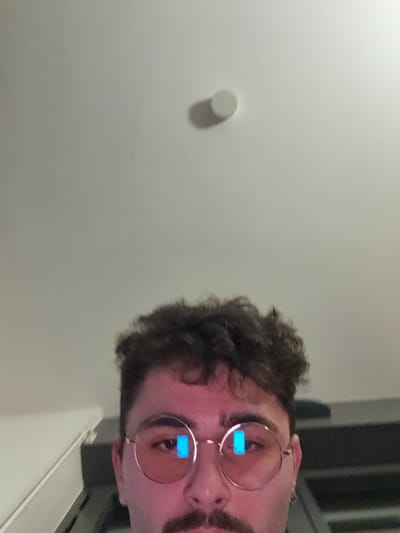 A man with curly brown hair, a mustache, and round spectacles looks at the camera. The light in the room is dim. A white wall with a small circular object sits to the left. A dark-colored object, potentially a frame of some kind, sits on the right, partially obscured. The image is oriented so the man is viewing from a sideways perspective.