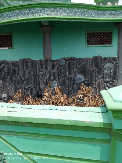 The image shows a close-up view of a section of a building in Kecamatan Haurgeulis, Indonesia. The building is painted a bright green and has a stone-like patterned facade. The facade is made up of small grey stones and features an intricate design of a mosque. Above the facade, a green decorative band lines the edge of the building. The building has a window on the right side of the image that has a decorative design and is also painted a vibrant green. There are dried plants visible beneath the facade. The image was taken at 12:44 PM on March 23, 2023.