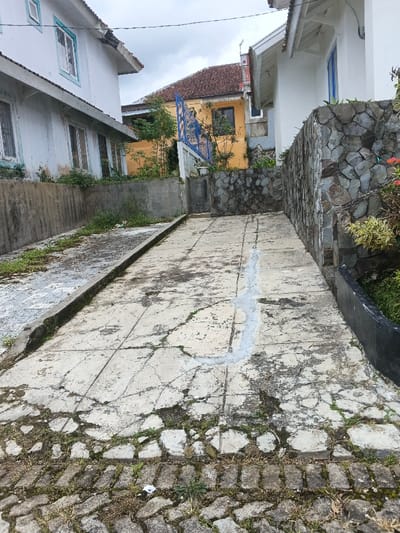 The image depicts an outdoor concrete pathway in the city of Pô Kô, Vietnam, flanked by residential structures and retaining walls under an overcast sky. The foreground features a section of dark gray paving stones with interspersed weeds, transitioning into a wider light gray concrete path. This concrete path is heavily cracked, stained, and overgrown with moss and weeds, and a prominent, winding light blue or white line of sealant or paint runs down its center.

On the left, a dark gray concrete retaining wall supports a multi-story white building with light blue window trim and a reddish-brown tiled roof. Vegetation grows over the top of this wall. On the right, a retaining wall constructed from irregular dark gray natural stones is visible, featuring small green plants and sparse red flowers along its top. This wall partially obscures another white building with a blue-framed window. In the background, a distinct yellow building with a reddish-brown tiled roof is visible, fronted by a blue, ornate metal gate or fence and green foliage. No individuals or active subjects are present in the scene.