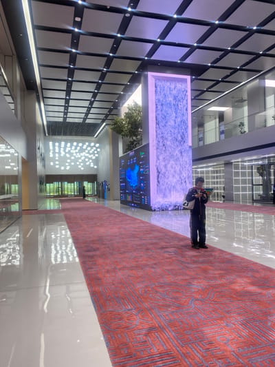 The image shows a large, modern, open interior space with a high ceiling and a tiled floor. The ceiling has a grid pattern of square tiles and recessed lighting. A red carpet runs down the center of the space, extending into the distance. A large digital screen on the right side of the image displays a complex map and data. Behind the screen is a wall with a tall, rectangular opening. The opening contains a large, shimmering, translucent waterfall. A young person wearing a blue jacket and black pants is standing on the carpet, looking down at a mobile phone in their left hand. They are standing to the right of the carpet, and to the left of the screen, directly in front of the waterfall. The person is facing the camera, but their face is obscured by their hair. The environment is clean and bright, with no other people present. The coordinates (latitude: 30.449, longitude: 120.242) place the location in the city of Hang Zhou Shi, China. The interior appears to be part of a large office building or other commercial space.